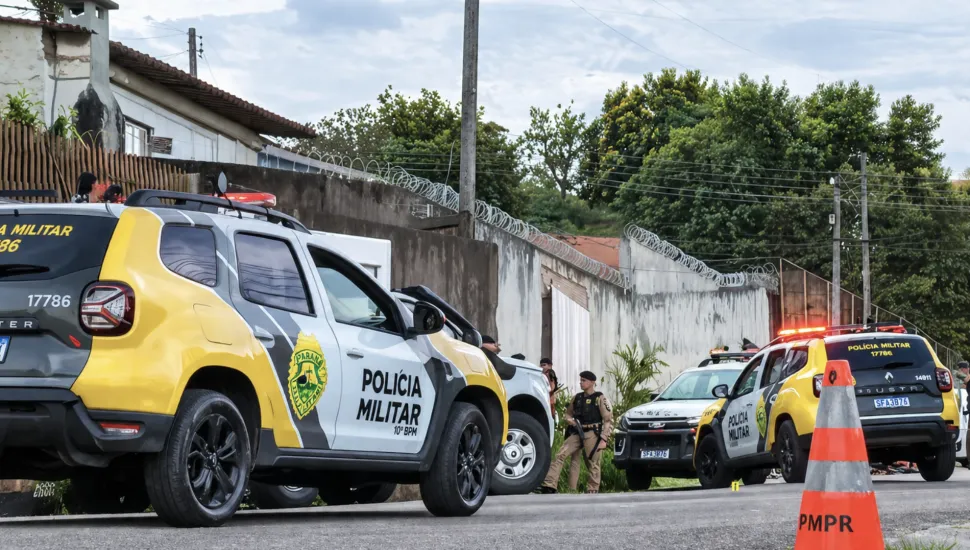 Fotografia em enquadramento médio e ângulo baixo de viaturas da Polícia Militar do Paraná estacionadas em uma rua residencial. Em primeiro plano, à esquerda, destaca-se a lateral de uma SUV amarela, cinza e branca com a inscrição "POLÍCIA MILITAR 10º BPM" e o brasão do estado na porta. Em segundo plano, no centro, um policial militar fardado e de boina preta está de pé ao lado de outro veículo policial. Ao fundo, à direita, outra viatura com os giroflex acesos em vermelho estaciona próximo a um muro alto com arame de proteção. Em primeiro plano, à extrema direita, há um cone de trânsito alaranjado com a sigla "PMPR".