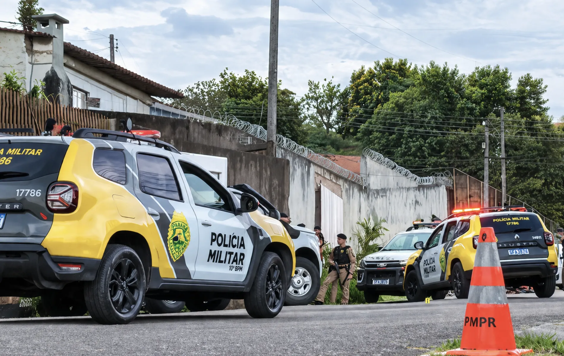 Fotografia em enquadramento médio e ângulo baixo de viaturas da Polícia Militar do Paraná estacionadas em uma rua residencial. Em primeiro plano, à esquerda, destaca-se a lateral de uma SUV amarela, cinza e branca com a inscrição "POLÍCIA MILITAR 10º BPM" e o brasão do estado na porta. Em segundo plano, no centro, um policial militar fardado e de boina preta está de pé ao lado de outro veículo policial. Ao fundo, à direita, outra viatura com os giroflex acesos em vermelho estaciona próximo a um muro alto com arame de proteção. Em primeiro plano, à extrema direita, há um cone de trânsito alaranjado com a sigla "PMPR".