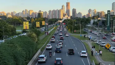 Fotografia em enquadramento amplo e ângulo elevado de uma grande avenida em Curitiba com tráfego intenso de veículos ao final da tarde. No centro, a via de múltiplas faixas exibe carros e motocicletas transitando em direção ao horizonte. À esquerda, estende-se uma ampla área verde arborizada com painéis publicitários amarelos e escuros ao longo do caminho. À direita, vê-se a calçada com pedestres caminhando, áreas de estacionamento e um totem vertical do ParkShoppingBarigüi. Ao fundo, a silhueta da cidade destaca diversos prédios residenciais e comerciais iluminados pela luz do pôr do sol.
