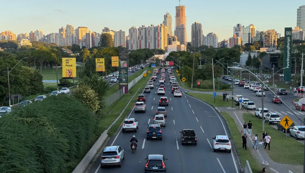 Fotografia em enquadramento amplo e ângulo elevado de uma grande avenida em Curitiba com tráfego intenso de veículos ao final da tarde. No centro, a via de múltiplas faixas exibe carros e motocicletas transitando em direção ao horizonte. À esquerda, estende-se uma ampla área verde arborizada com painéis publicitários amarelos e escuros ao longo do caminho. À direita, vê-se a calçada com pedestres caminhando, áreas de estacionamento e um totem vertical do ParkShoppingBarigüi. Ao fundo, a silhueta da cidade destaca diversos prédios residenciais e comerciais iluminados pela luz do pôr do sol.