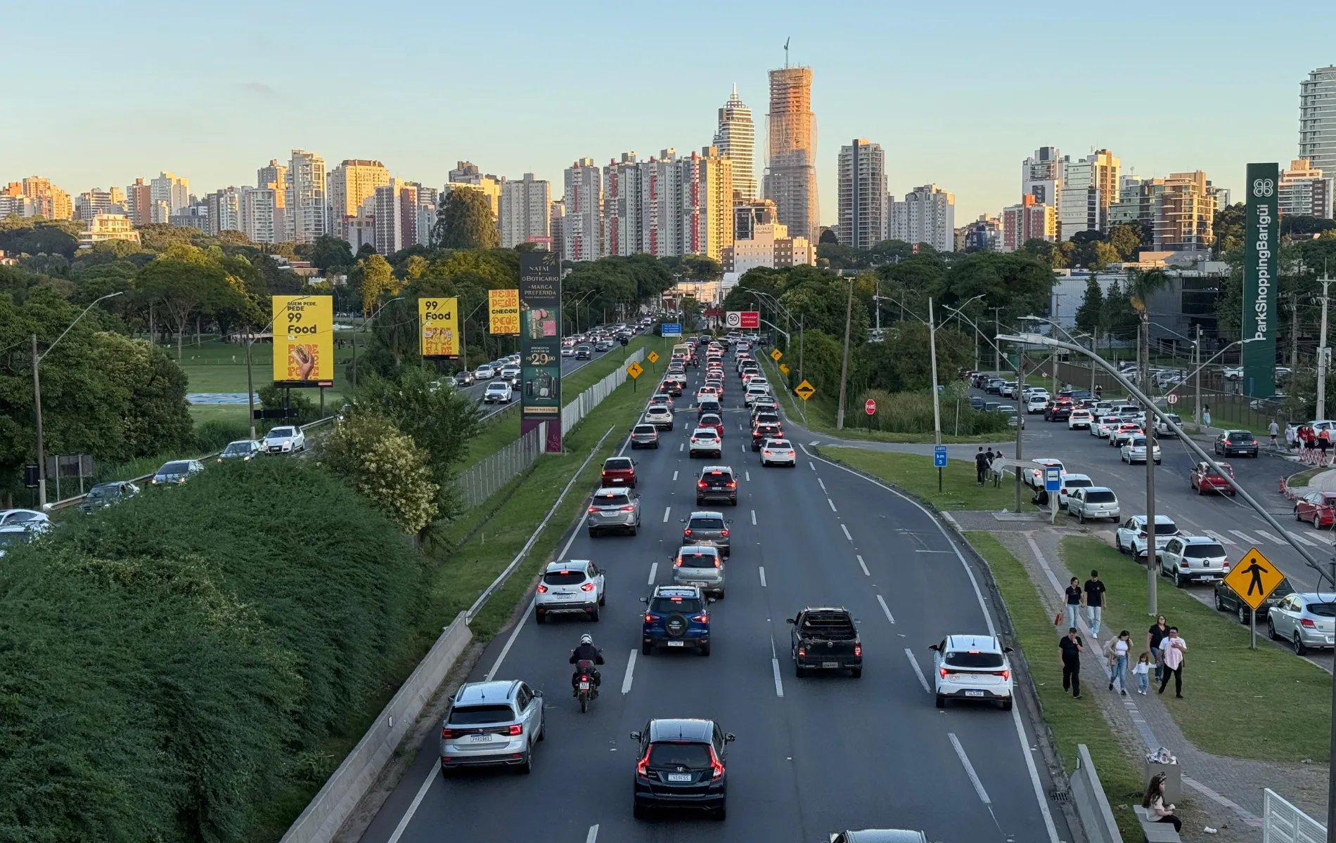 Fotografia em enquadramento amplo e ângulo elevado de uma grande avenida em Curitiba com tráfego intenso de veículos ao final da tarde. No centro, a via de múltiplas faixas exibe carros e motocicletas transitando em direção ao horizonte. À esquerda, estende-se uma ampla área verde arborizada com painéis publicitários amarelos e escuros ao longo do caminho. À direita, vê-se a calçada com pedestres caminhando, áreas de estacionamento e um totem vertical do ParkShoppingBarigüi. Ao fundo, a silhueta da cidade destaca diversos prédios residenciais e comerciais iluminados pela luz do pôr do sol.