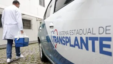 Imagem mostra homem vestido de branco carregando um cooler, saino de um carro que na porta esta escrito Sistema Estadual de Transplantes