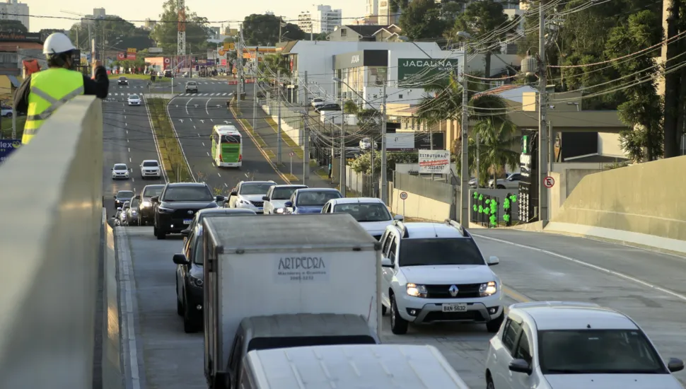 Fotografia em enquadramento amplo e plano profundo de uma via urbana recém-construída ou reformada com tráfego de veículos. Em primeiro plano, no canto superior esquerdo, um trabalhador vestindo colete refletivo amarelo e capacete branco observa o trânsito sobre a mureta de contenção de uma estrutura elevada. Abaixo, carros e caminhões baú transitam pelas pistas pavimentadas de concreto. Ao longo da via, veem-se canteiros, postes com fiação elétrica, imóveis comerciais, vegetação urbana e edifícios ao fundo sob iluminação solar diurna.