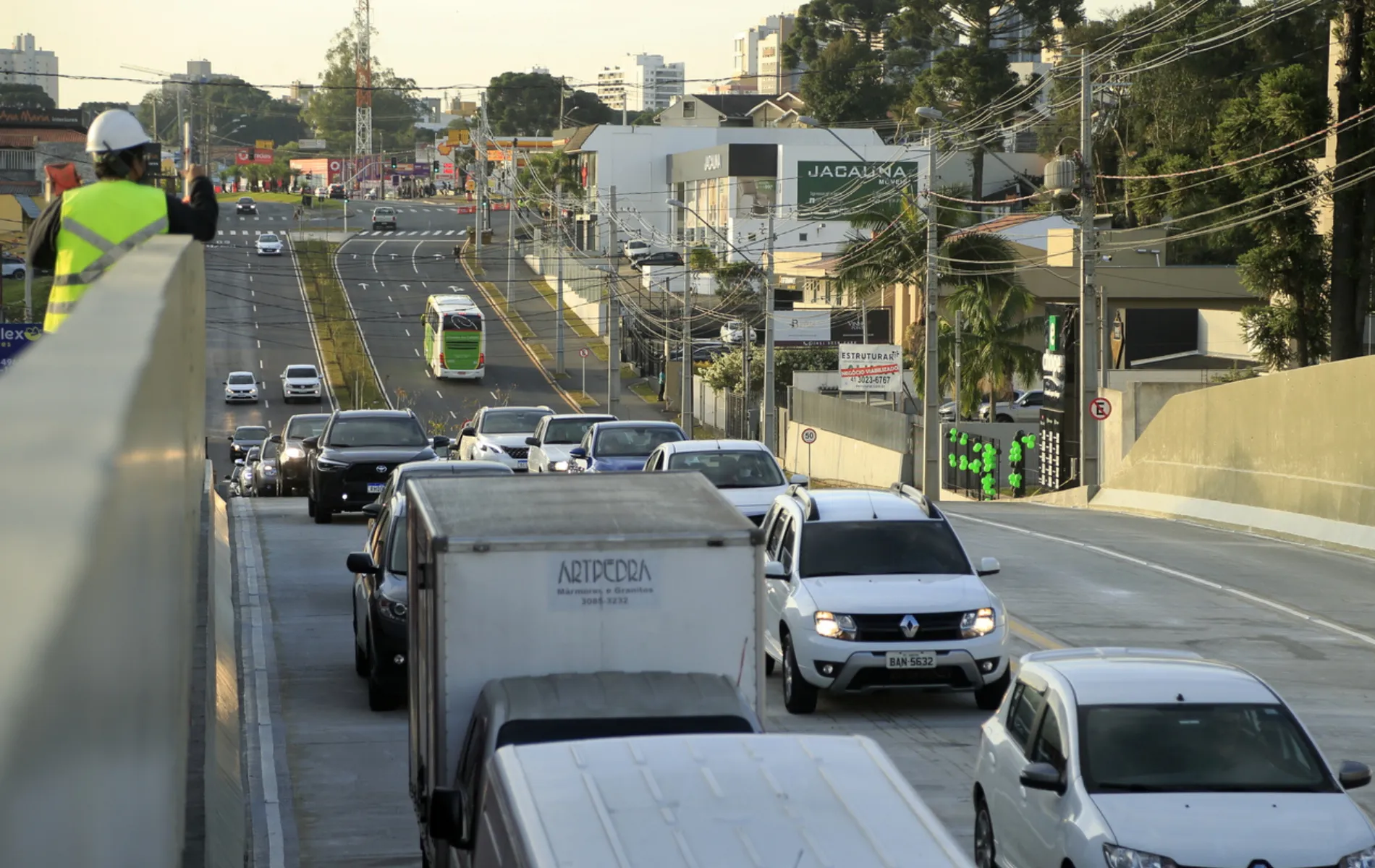 Fotografia em enquadramento amplo e plano profundo de uma via urbana recém-construída ou reformada com tráfego de veículos. Em primeiro plano, no canto superior esquerdo, um trabalhador vestindo colete refletivo amarelo e capacete branco observa o trânsito sobre a mureta de contenção de uma estrutura elevada. Abaixo, carros e caminhões baú transitam pelas pistas pavimentadas de concreto. Ao longo da via, veem-se canteiros, postes com fiação elétrica, imóveis comerciais, vegetação urbana e edifícios ao fundo sob iluminação solar diurna.