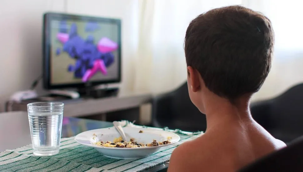 Imagem mostra o uso de tela durante uma refeição: um menino de costas com o prato cheio, como cheio e assistindo a um desenho.