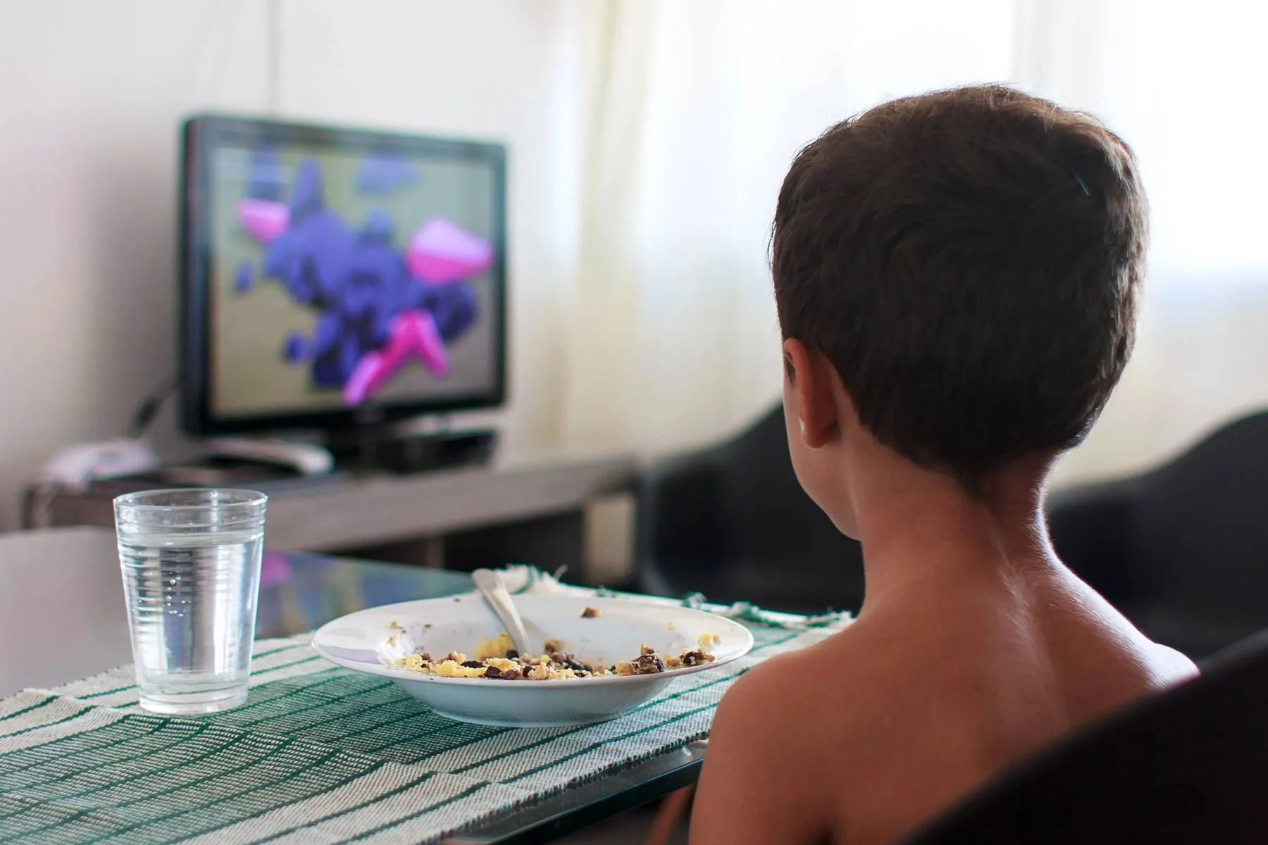 Imagem mostra o uso de tela durante uma refeição: um menino de costas com o prato cheio, como cheio e assistindo a um desenho.