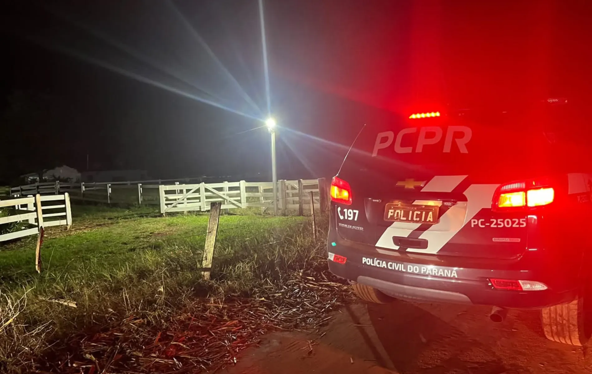 Fotografia em enquadramento médio e visão traseira de uma viatura da Polícia Civil do Paraná durante uma operação noturna em área rural. À direita, sobressai-se a traseira do veículo caracterizado com a sigla "PCPR", a inscrição "POLÍCIA CIVIL DO PARANÁ", o número de emergência "197" e lanternas vermelhas acesas lançando forte brilho no ambiente. À esquerda, em meio à escuridão da noite iluminada por um ponto de luz ao fundo, veem-se Cercas de madeira branca delimitando uma propriedade rural e vegetação rasteira ao longo da estrada de terra.