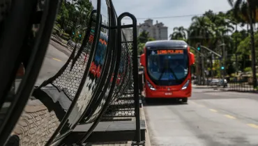 Fotografia em enquadramento médio com foco seletivo no sistema de transporte coletivo de Curitiba. Em primeiro plano, à esquerda, sobressai-se a estrutura cilíndrica de vidro de uma estação-tubo, refletindo a calçada em petit-pavé e a vegetação do entorno. Em segundo plano, no lado direito, um ônibus biarticulado vermelho aproxima-se pela canaleta exclusiva com o letreiro luminoso laranja aceso. Ao fundo, veem-se semáforos, árvores e edifícios urbanos sob iluminação diurna.