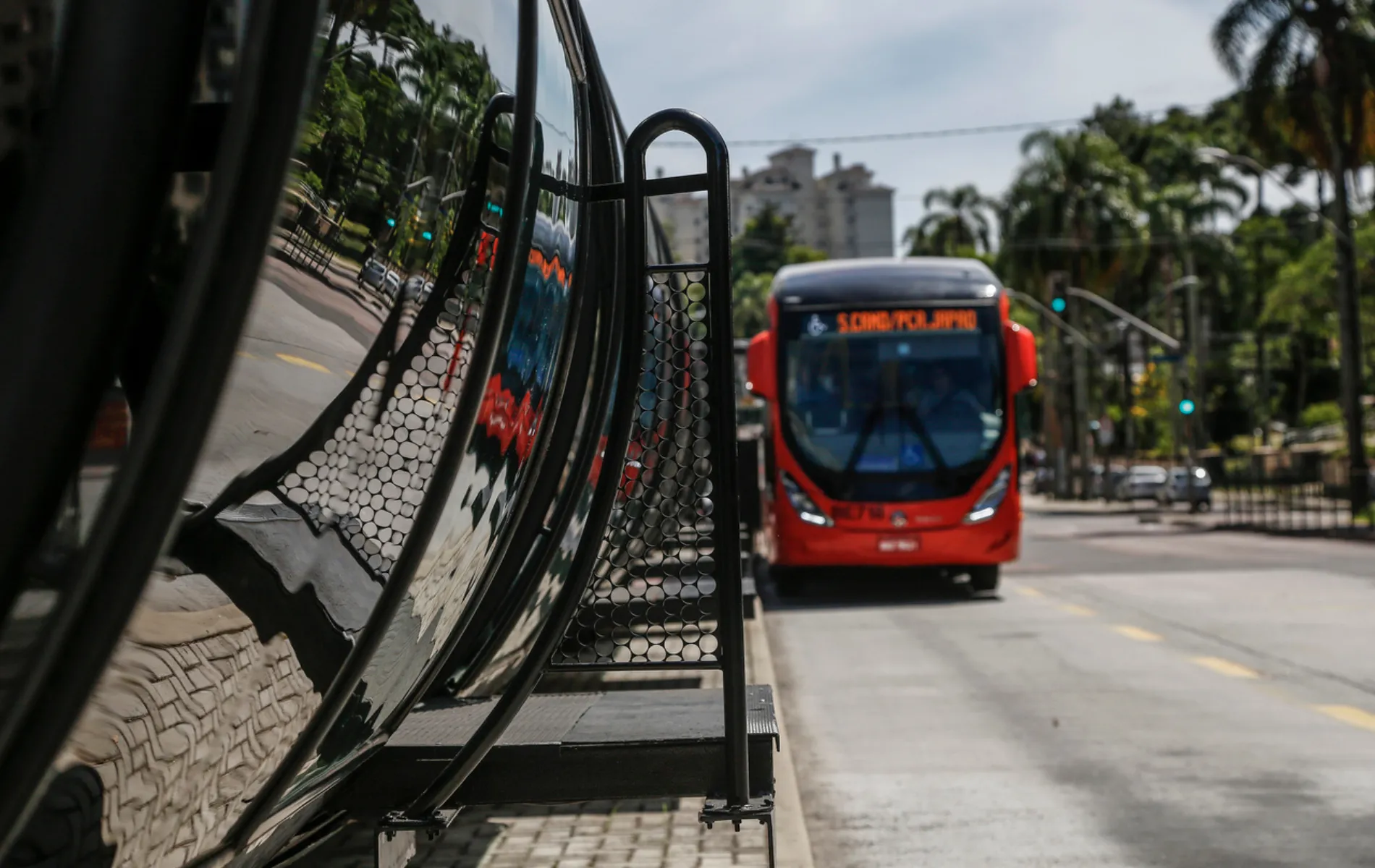 Fotografia em enquadramento médio com foco seletivo no sistema de transporte coletivo de Curitiba. Em primeiro plano, à esquerda, sobressai-se a estrutura cilíndrica de vidro de uma estação-tubo, refletindo a calçada em petit-pavé e a vegetação do entorno. Em segundo plano, no lado direito, um ônibus biarticulado vermelho aproxima-se pela canaleta exclusiva com o letreiro luminoso laranja aceso. Ao fundo, veem-se semáforos, árvores e edifícios urbanos sob iluminação diurna.