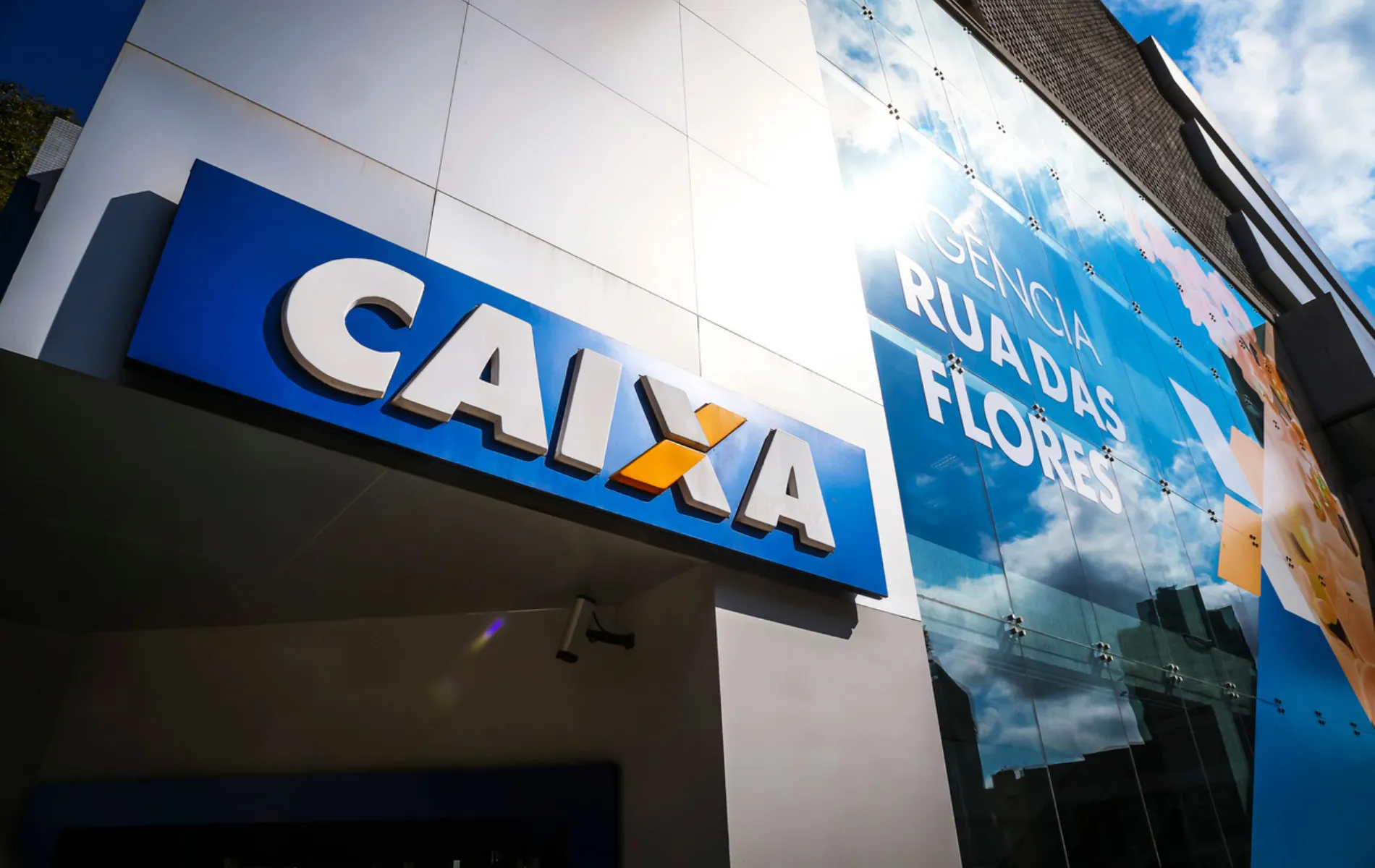 Fotografia em enquadramento aproximado e ângulo em contra-plongée da fachada de uma agência bancária da Caixa Econômica Federal. Em primeiro plano, à esquerda, destaca-se um letreiro retangular azul sob a parede branca com a palavra "CAIXA" grafada em letras brancas tridimensionais, com o detalhe do "X" estilizado em laranja. À direita, a fachada espelhada em painéis de vidro reflete o céu azul com nuvens e o brilho intenso do sol, além de exibir a inscrição adesivada "AGÊNCIA RUA DAS FLORES".