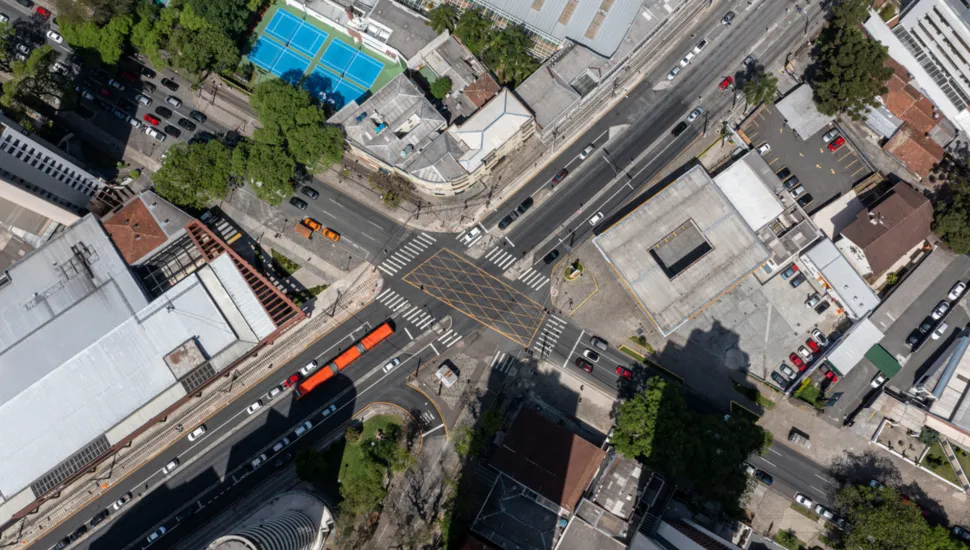 Fotografia em vista aérea superior de um cruzamento viário urbano em área adensada. Ao centro, destaca-se a interseção das vias marcada com sinalização horizontal amarela em formato de quadriculado no asfalto (área de conflito) e faixas de pedestres nos quatro acessos. Pistas de rolamento cortam a imagem na diagonal, onde transitam veículos de passeio e um ônibus biarticulado vermelho. No entorno do cruzamento, distribuem-se quadras com edifícios residenciais e comerciais, telhados diversos, pátios de estacionamento, quadras esportivas de piso azul e copas de árvores que projetam sombras sobre as vias sob luz solar direta.