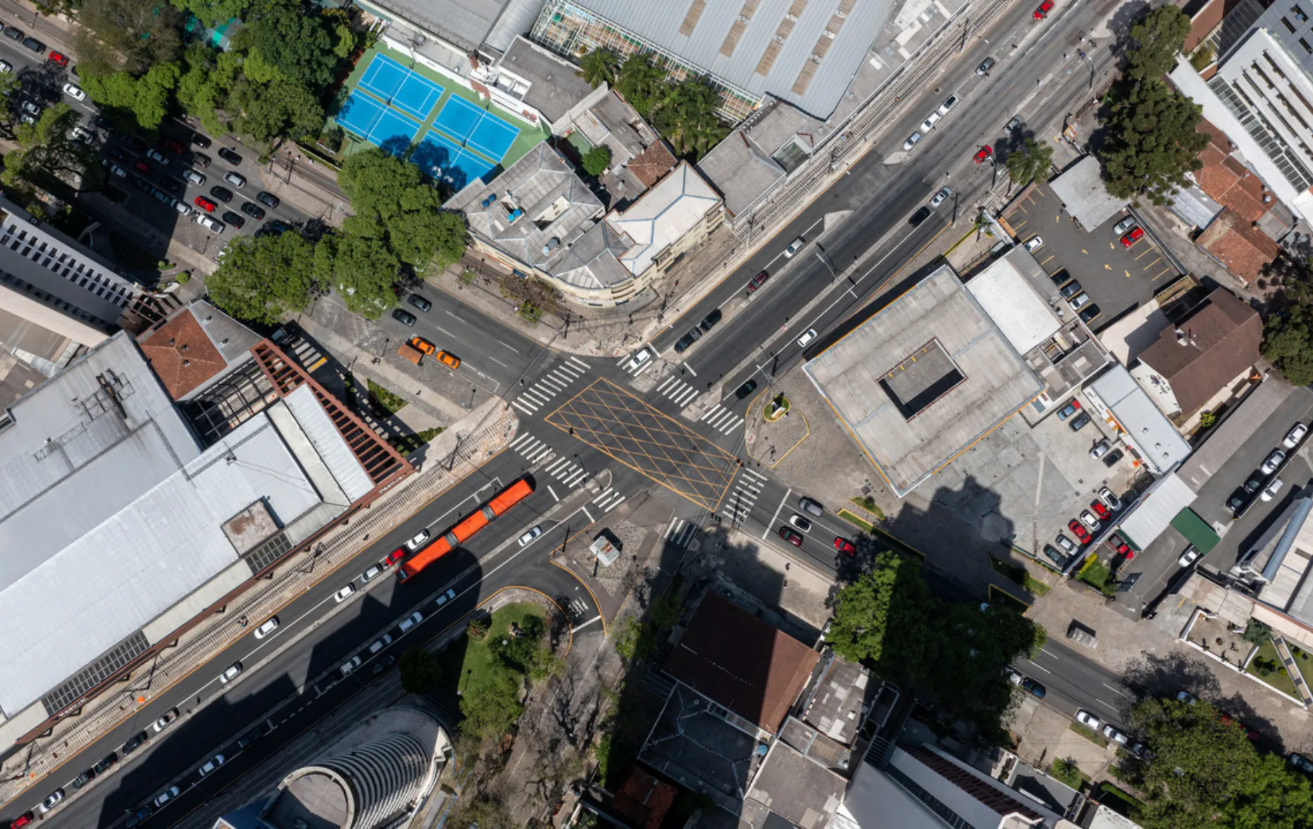 Fotografia em vista aérea superior de um cruzamento viário urbano em área adensada. Ao centro, destaca-se a interseção das vias marcada com sinalização horizontal amarela em formato de quadriculado no asfalto (área de conflito) e faixas de pedestres nos quatro acessos. Pistas de rolamento cortam a imagem na diagonal, onde transitam veículos de passeio e um ônibus biarticulado vermelho. No entorno do cruzamento, distribuem-se quadras com edifícios residenciais e comerciais, telhados diversos, pátios de estacionamento, quadras esportivas de piso azul e copas de árvores que projetam sombras sobre as vias sob luz solar direta.