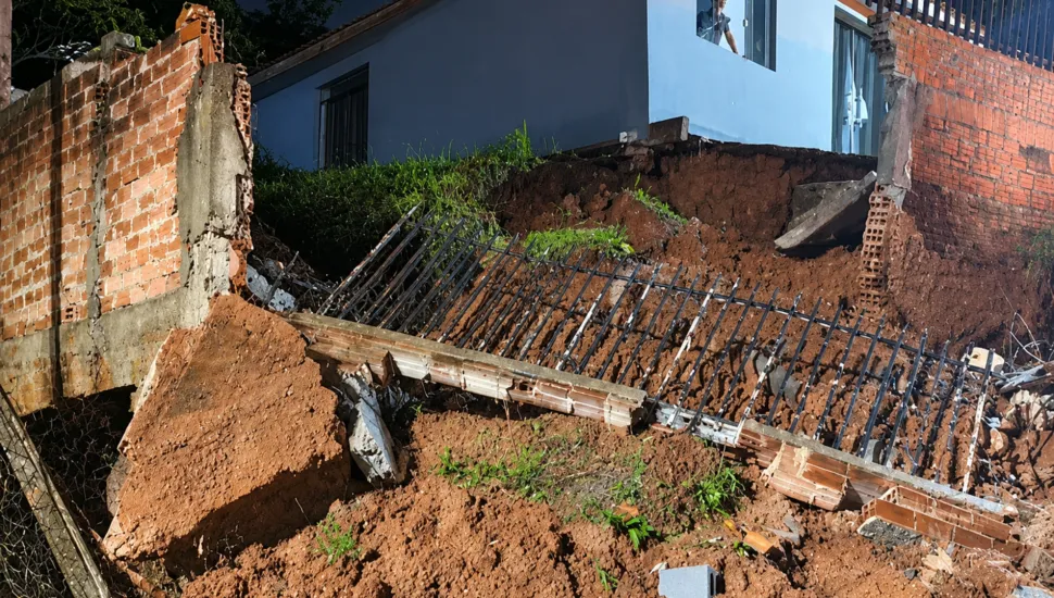 Fotografia em enquadramento médio mostrando os estragos de um desmoronamento de terra em uma área residencial. No centro da imagem, uma grande quantidade de terra avermelhada cedeu, derrubando uma estrutura de muro e o gradil metálico preto que ficava no topo, o qual jaz caído e inclinado sobre o barranco. À esquerda e à direita, veem-se os trechos remanescentes do muro de tijolos alaranjados expostos. Ao fundo, em nível elevado em relação ao solo desabado, fica uma casa de parede azul e, da janela superior, uma pessoa observa os danos causados pelo deslizamento.