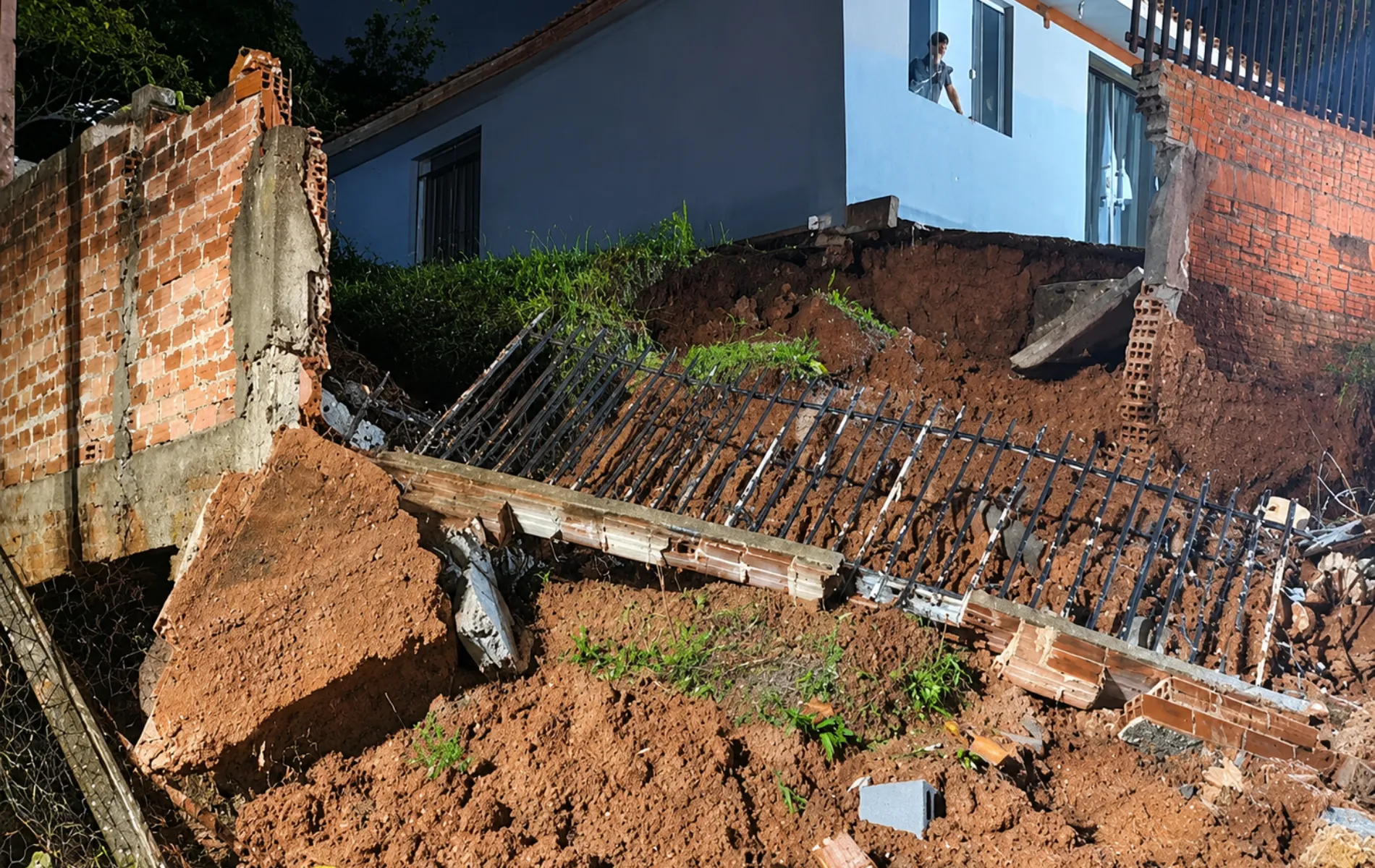 Fotografia em enquadramento médio mostrando os estragos de um desmoronamento de terra em uma área residencial. No centro da imagem, uma grande quantidade de terra avermelhada cedeu, derrubando uma estrutura de muro e o gradil metálico preto que ficava no topo, o qual jaz caído e inclinado sobre o barranco. À esquerda e à direita, veem-se os trechos remanescentes do muro de tijolos alaranjados expostos. Ao fundo, em nível elevado em relação ao solo desabado, fica uma casa de parede azul e, da janela superior, uma pessoa observa os danos causados pelo deslizamento.