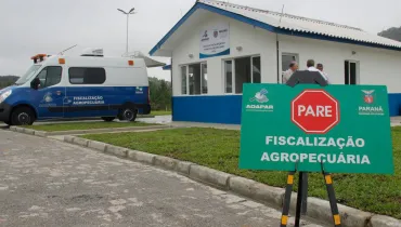 Fotografia em enquadramento médio e ângulo baixo de um posto de fiscalização agropecuária. Em primeiro plano, à direita, destaca-se uma placa verde sobre cavalete com o símbolo de um octógono vermelho com a palavra "PARE", além do logotipo da Adapar, o brasão do Governo do Paraná e o texto "FISCALIZAÇÃO AGROPECUÁRIA" em letras brancas. Ao fundo, no centro, vê-se um prédio térreo de paredes brancas com base azul e telhado em duas águas, onde três pessoas conversam na entrada. À esquerda, estacionada no pátio, fica uma furgoneta caracterizada da fiscalização agropecuária sob iluminação natural de dia nublado.