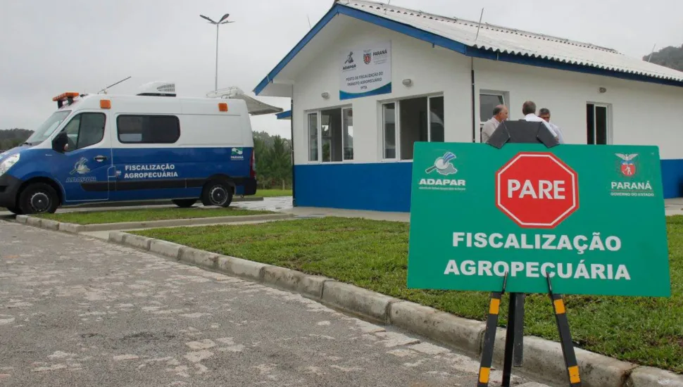 Fotografia em enquadramento médio e ângulo baixo de um posto de fiscalização agropecuária. Em primeiro plano, à direita, destaca-se uma placa verde sobre cavalete com o símbolo de um octógono vermelho com a palavra "PARE", além do logotipo da Adapar, o brasão do Governo do Paraná e o texto "FISCALIZAÇÃO AGROPECUÁRIA" em letras brancas. Ao fundo, no centro, vê-se um prédio térreo de paredes brancas com base azul e telhado em duas águas, onde três pessoas conversam na entrada. À esquerda, estacionada no pátio, fica uma furgoneta caracterizada da fiscalização agropecuária sob iluminação natural de dia nublado.