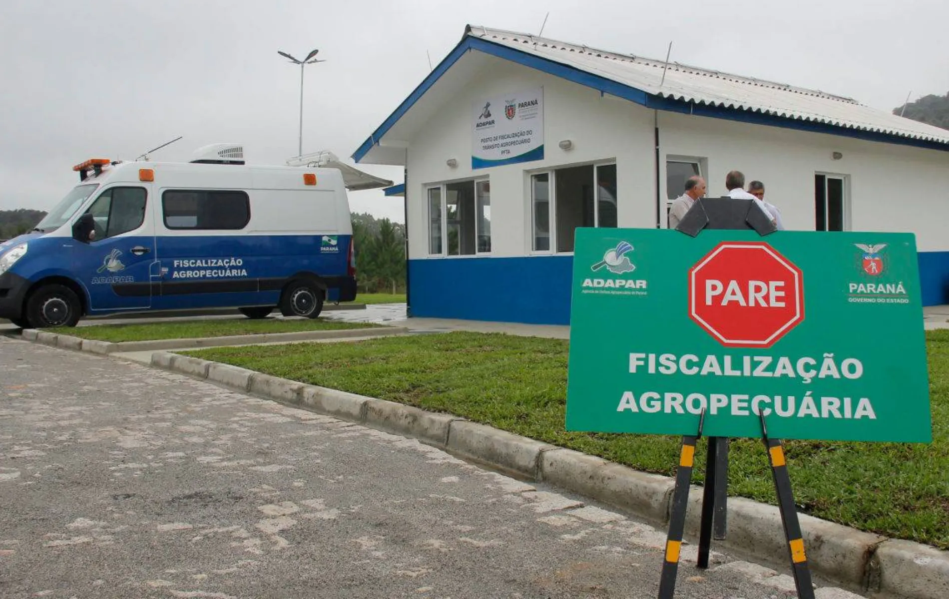Fotografia em enquadramento médio e ângulo baixo de um posto de fiscalização agropecuária. Em primeiro plano, à direita, destaca-se uma placa verde sobre cavalete com o símbolo de um octógono vermelho com a palavra "PARE", além do logotipo da Adapar, o brasão do Governo do Paraná e o texto "FISCALIZAÇÃO AGROPECUÁRIA" em letras brancas. Ao fundo, no centro, vê-se um prédio térreo de paredes brancas com base azul e telhado em duas águas, onde três pessoas conversam na entrada. À esquerda, estacionada no pátio, fica uma furgoneta caracterizada da fiscalização agropecuária sob iluminação natural de dia nublado.