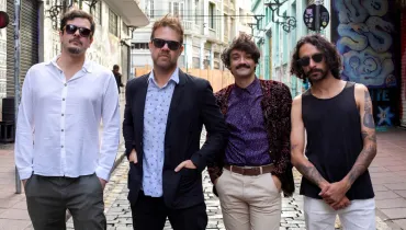 Fotografia em enquadramento médio e plano conjunto de quatro homens posicionados lado a lado em uma rua de paralelepípedos. Da esquerda para a direita: O primeiro homem usa óculos escuros, camisa social branca de mangas compridas e calça cinza; o segundo veste blazer escuro sobre camisa social clara estampa de bolinhas e óculos escuros; o terceiro tem bigode, cabelo ondulado e veste camisa roxa com blazer aveludado estampado e calça clara; o quarto homem possui cabelos cacheados na altura dos ombros, óculos escuros, regata preta que deixa à mostra braços tatuados e calça branca.
