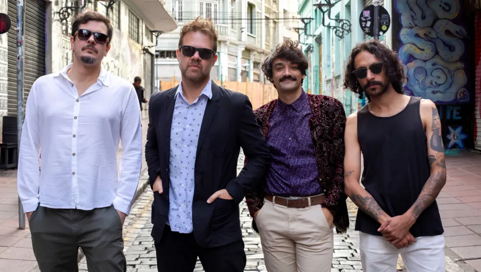 Fotografia em enquadramento médio e plano conjunto de quatro homens posicionados lado a lado em uma rua de paralelepípedos. Da esquerda para a direita: O primeiro homem usa óculos escuros, camisa social branca de mangas compridas e calça cinza; o segundo veste blazer escuro sobre camisa social clara estampa de bolinhas e óculos escuros; o terceiro tem bigode, cabelo ondulado e veste camisa roxa com blazer aveludado estampado e calça clara; o quarto homem possui cabelos cacheados na altura dos ombros, óculos escuros, regata preta que deixa à mostra braços tatuados e calça branca.