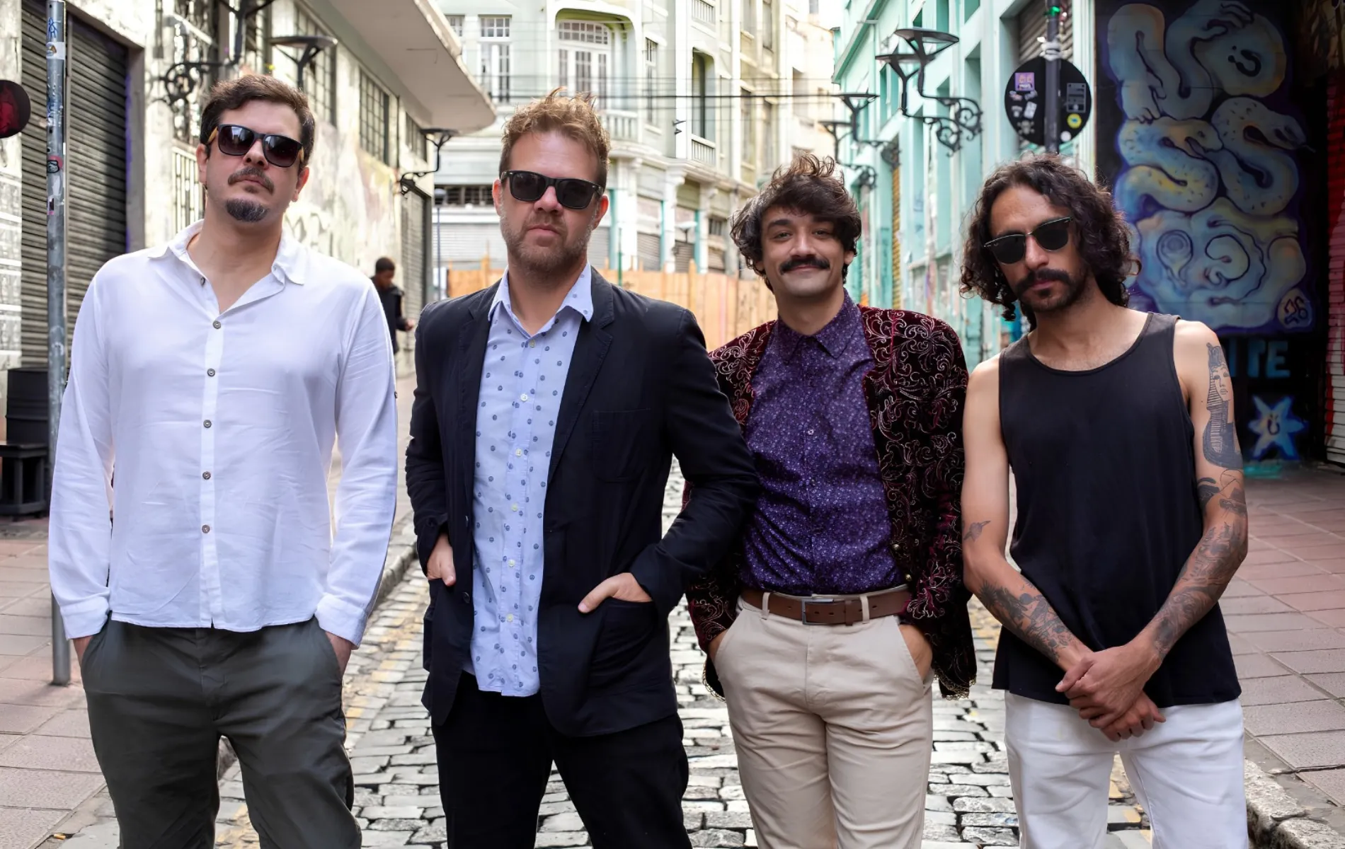 Fotografia em enquadramento médio e plano conjunto de quatro homens posicionados lado a lado em uma rua de paralelepípedos. Da esquerda para a direita: O primeiro homem usa óculos escuros, camisa social branca de mangas compridas e calça cinza; o segundo veste blazer escuro sobre camisa social clara estampa de bolinhas e óculos escuros; o terceiro tem bigode, cabelo ondulado e veste camisa roxa com blazer aveludado estampado e calça clara; o quarto homem possui cabelos cacheados na altura dos ombros, óculos escuros, regata preta que deixa à mostra braços tatuados e calça branca.