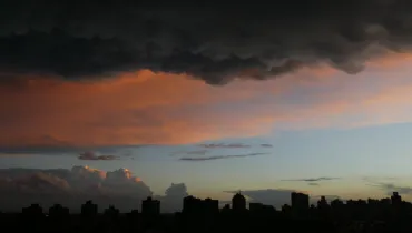 Fotografia em enquadramento amplo do horizonte urbano durante o crepúsculo. Na metade inferior, a silhueta escura de diversos edifícios da cidade desenha a linha do horizonte contra o céu. O céu apresenta um gradiente de cores que varia do azul-claro próximo ao horizonte até tons alaranjados e rosados no centro, causados pela iluminação do pôr do sol. Na parte superior, densas nuvens escuras de tempestade cobrem o topo da composição, criando um forte contraste de iluminação e cor sobre a paisagem.