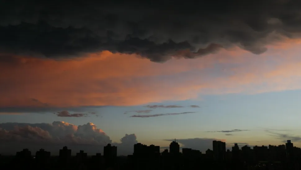 Fotografia em enquadramento amplo do horizonte urbano durante o crepúsculo. Na metade inferior, a silhueta escura de diversos edifícios da cidade desenha a linha do horizonte contra o céu. O céu apresenta um gradiente de cores que varia do azul-claro próximo ao horizonte até tons alaranjados e rosados no centro, causados pela iluminação do pôr do sol. Na parte superior, densas nuvens escuras de tempestade cobrem o topo da composição, criando um forte contraste de iluminação e cor sobre a paisagem.