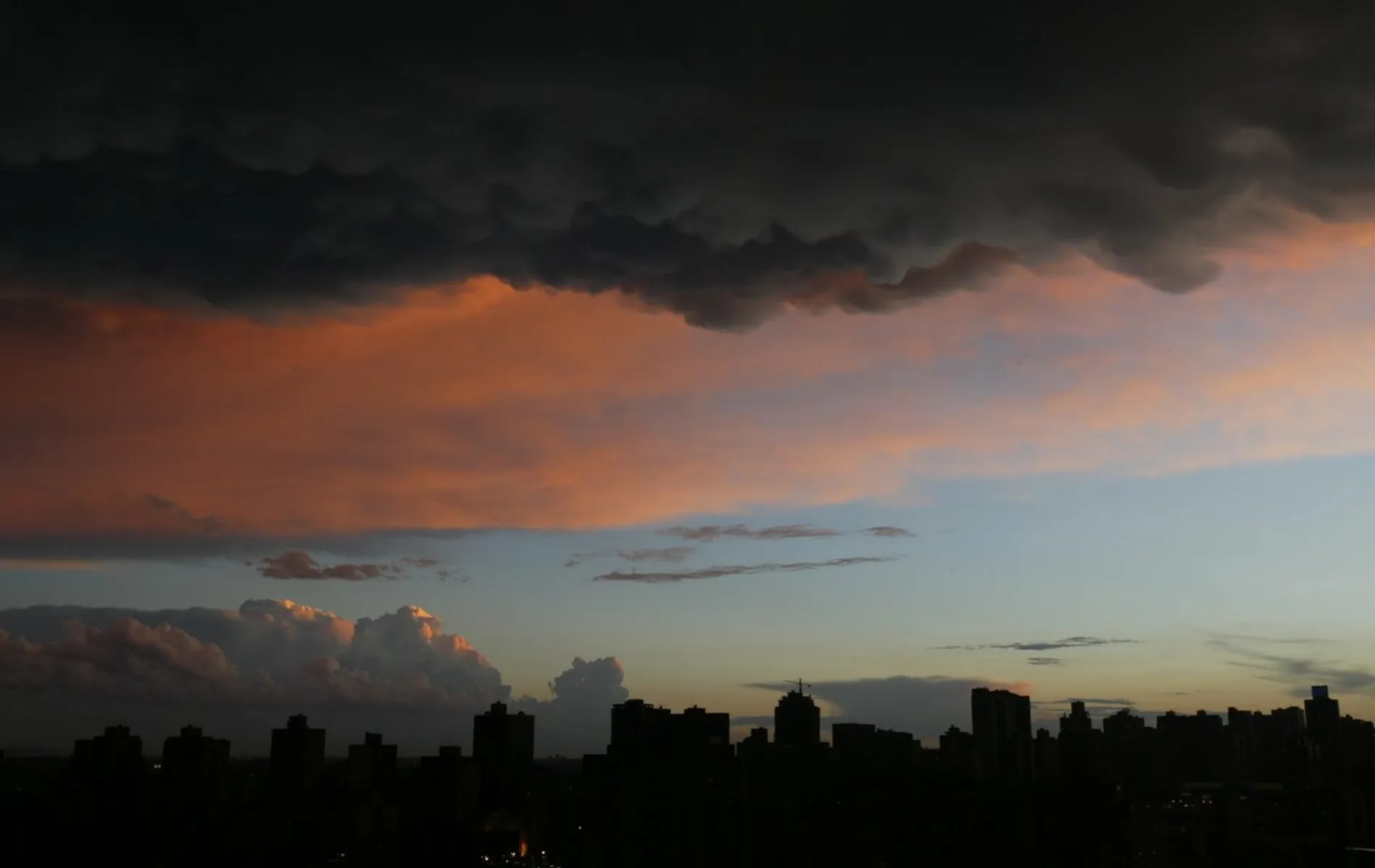 Fotografia em enquadramento amplo do horizonte urbano durante o crepúsculo. Na metade inferior, a silhueta escura de diversos edifícios da cidade desenha a linha do horizonte contra o céu. O céu apresenta um gradiente de cores que varia do azul-claro próximo ao horizonte até tons alaranjados e rosados no centro, causados pela iluminação do pôr do sol. Na parte superior, densas nuvens escuras de tempestade cobrem o topo da composição, criando um forte contraste de iluminação e cor sobre a paisagem.