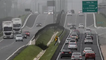 Fotografia em enquadramento amplo e perspectiva longitudinal de uma rodovia movimentada em um dia com forte neblina. A pista divide-se em dois sentidos, separados por um canteiro central gramado. À esquerda, caminhões brancos com a inscrição "SULISTA" e carros transitam sob a bruma densa. À direita, o tráfego de veículos de passeio e utilitários é mais intenso, seguindo em direção a uma elevação da pista. Acima da pista da direita, vê-se uma placa verde de sinalização viária com a indicação "SAÍDA 74 - RETORNO - S. J. dos Pinhais - Piraquara". Ao fundo, a paisagem e a fiação elétrica perdem visibilidade devido ao nevoeiro denso.