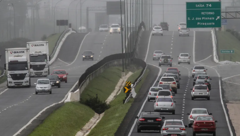 Fotografia em enquadramento amplo e perspectiva longitudinal de uma rodovia movimentada em um dia com forte neblina. A pista divide-se em dois sentidos, separados por um canteiro central gramado. À esquerda, caminhões brancos com a inscrição "SULISTA" e carros transitam sob a bruma densa. À direita, o tráfego de veículos de passeio e utilitários é mais intenso, seguindo em direção a uma elevação da pista. Acima da pista da direita, vê-se uma placa verde de sinalização viária com a indicação "SAÍDA 74 - RETORNO - S. J. dos Pinhais - Piraquara". Ao fundo, a paisagem e a fiação elétrica perdem visibilidade devido ao nevoeiro denso.