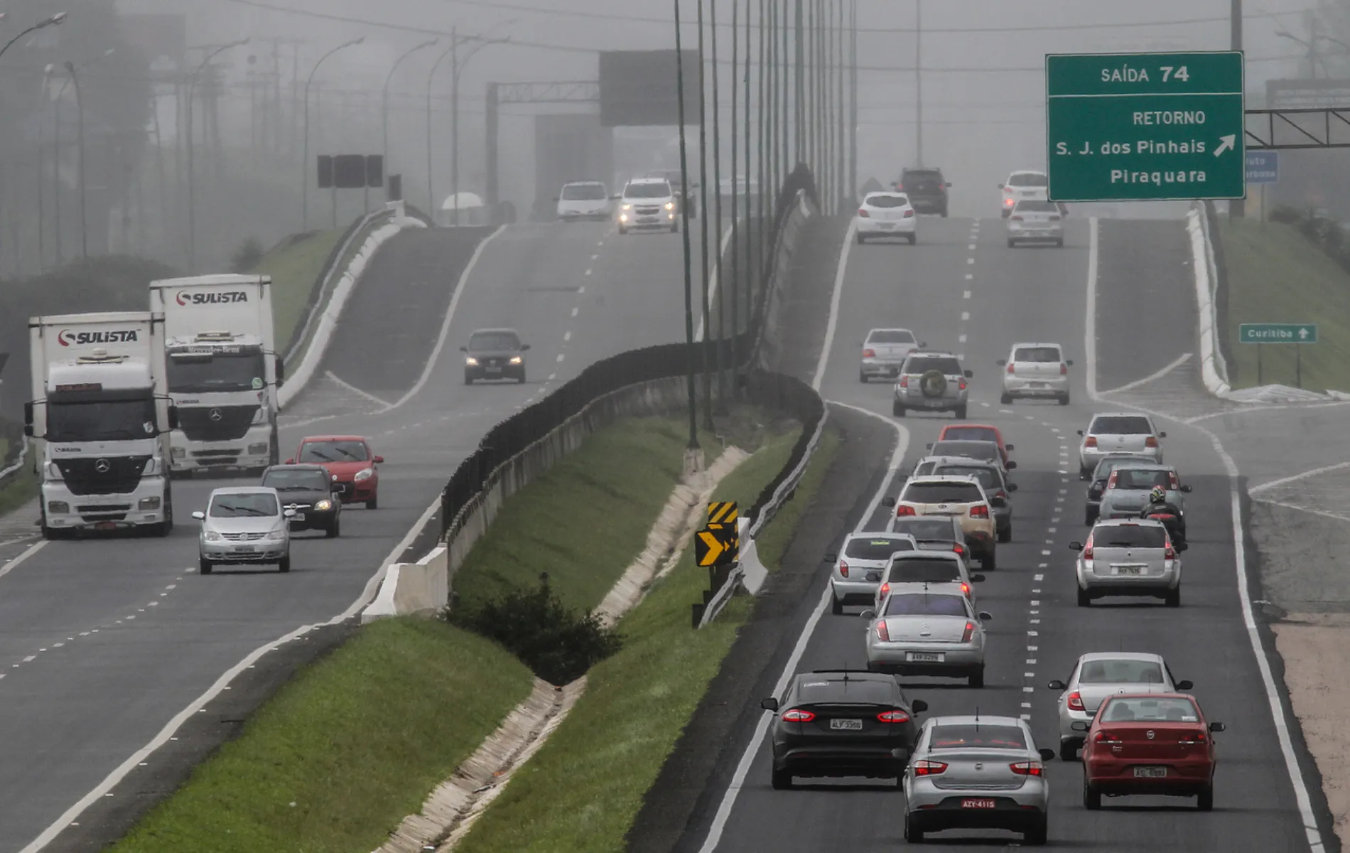 Fotografia em enquadramento amplo e perspectiva longitudinal de uma rodovia movimentada em um dia com forte neblina. A pista divide-se em dois sentidos, separados por um canteiro central gramado. À esquerda, caminhões brancos com a inscrição "SULISTA" e carros transitam sob a bruma densa. À direita, o tráfego de veículos de passeio e utilitários é mais intenso, seguindo em direção a uma elevação da pista. Acima da pista da direita, vê-se uma placa verde de sinalização viária com a indicação "SAÍDA 74 - RETORNO - S. J. dos Pinhais - Piraquara". Ao fundo, a paisagem e a fiação elétrica perdem visibilidade devido ao nevoeiro denso.