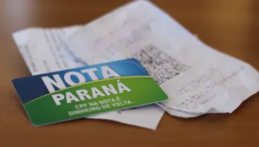 #PraCegoVer #DescriçãoDaImagem Fotografia em enquadramento aproximado e ângulo plongée de um cartão do programa Nota Paraná repousando sobre cupom fiscal e notas fiscais de papel. Em primeiro plano, no canto inferior esquerdo, destaca-se o cartão magnético com design dividido em azul e verde, exibindo em letras brancas o texto "NOTA PARANÁ" e o slogan "CPF NA NOTA É DINHEIRO DE VOLTA.". Em segundo plano, ligeiramente desfocado, vê-se um comprovante fiscal impresso com um código QR impresso na parte central. Os itens estão sobre uma superfície de madeira marrom clara sob luz suave.