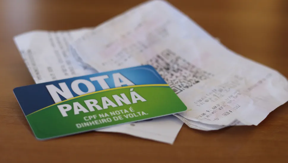 #PraCegoVer #DescriçãoDaImagem Fotografia em enquadramento aproximado e ângulo plongée de um cartão do programa Nota Paraná repousando sobre cupom fiscal e notas fiscais de papel. Em primeiro plano, no canto inferior esquerdo, destaca-se o cartão magnético com design dividido em azul e verde, exibindo em letras brancas o texto "NOTA PARANÁ" e o slogan "CPF NA NOTA É DINHEIRO DE VOLTA.". Em segundo plano, ligeiramente desfocado, vê-se um comprovante fiscal impresso com um código QR impresso na parte central. Os itens estão sobre uma superfície de madeira marrom clara sob luz suave.