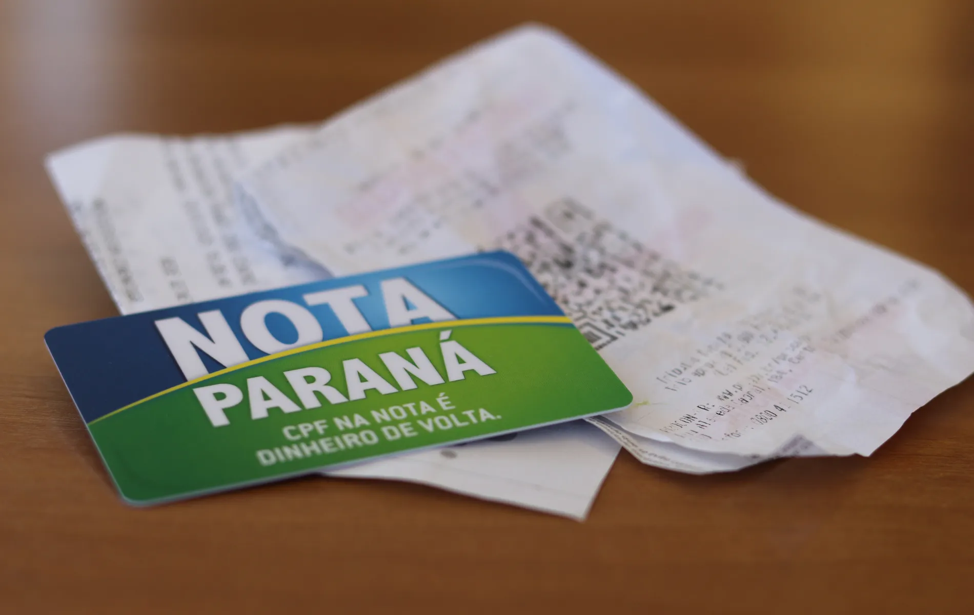 #PraCegoVer #DescriçãoDaImagem Fotografia em enquadramento aproximado e ângulo plongée de um cartão do programa Nota Paraná repousando sobre cupom fiscal e notas fiscais de papel. Em primeiro plano, no canto inferior esquerdo, destaca-se o cartão magnético com design dividido em azul e verde, exibindo em letras brancas o texto "NOTA PARANÁ" e o slogan "CPF NA NOTA É DINHEIRO DE VOLTA.". Em segundo plano, ligeiramente desfocado, vê-se um comprovante fiscal impresso com um código QR impresso na parte central. Os itens estão sobre uma superfície de madeira marrom clara sob luz suave.