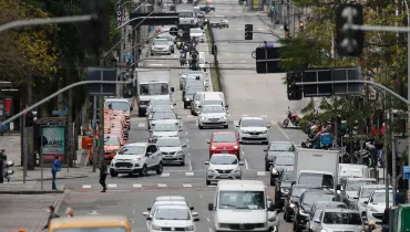 Fotografia em enquadramento amplo com plano de profundidade e teleobjetiva de uma avenida urbana movimentada. A via estende-se ao longo do quadro tomada por intenso fluxo de veículos, incluindo carros de passeio, furgões brancos, utilitários, motocicletas e uma fila de táxis laranjas estacionados ou em faixa dedicada à esquerda. Em primeiro plano, semáforos suspensos em hastes metálicas atravessam a parte superior da composição. Ao longo do canteiro e das calçadas, veem-se pedestres, fiação aérea emaranhada, pontos de ônibus e a arborização urbana sob luz natural de um dia nublado.