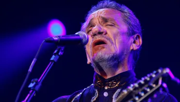 Fotografia em primeiro plano aproximado (close-up) e ângulo em contra-plongée do cantor e compositor Zé Ramalho durante uma apresentação musical. Ele está posicionado no centro do quadro, com a cabeça levemente inclinada para cima e de olhos fechados, expressando emoção ao cantar em um microfone posicionado à sua esquerda. Ele tem pele clara, cabelos e barba grisalhos com cavanhaque, e veste uma camisa escura com bordados dourados no colarinho. No canto inferior direito, vê-se a mão do instrumento musical que ele toca. O fundo é escuro, iluminado por refletores de palco em tons de roxo e azul, destacando o contorno do artista.