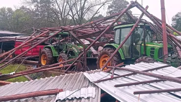 Fotografia em plano médio mostrando os estragos causados pelo desabamento da estrutura de um galpão rural. No centro da composição, sob a estrutura colapsada de caibros de madeira e vigas metálicas contorcidas, estão parcialmente soterrados dois tratores agrícolas na cor verde com rodas amarelas. Em primeiro plano, no canto inferior, veem-se chapas metálicas onduladas e prateadas do telhado arrancado e espalhado pelo chão. Ao fundo, contornando a cena sob céu nublado, observa-se uma linha de árvores de copa alta.