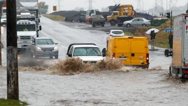 Fotografia em enquadramento amplo de um trecho urbano e rodoviário alagado durante uma chuva intensa. Em primeiro plano, a água barrenta cobre totalmente o asfalto, formando grandes marolas e respingos à medida que os veículos transitam. No centro, uma caminhonete branca com sanantonio e um furgão amarelo avançam pela alagação. À esquerda, outros carros e um caminhão branco com a inscrição "Mercedes-Benz" trafegam pela pista parcialmente coberta de água. À direita, pedestres com guarda-chuvas observam a movimentação na calçada. Ao fundo, veem-se máquinas pesadas, caminhões de carga e torres de alta tensão sob céu encoberto.