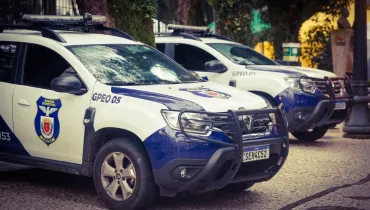 Fotografia em enquadramento médio e ângulo oblíquo de duas viaturas da Guarda Municipal de Curitiba estacionadas em uma praça com calçamento de pedras portuguesas. Em primeiro plano, à esquerda, destaca-se a parte dianteira e lateral de um utilitário esportivo (SUV) modelo Renault Duster, pintado nas cores branca e azul. A porta exibe o brasão da "Guarda Municipal Curitiba" e a identificação "GPEO 05" na lateral do capô, além de para-choque de impulsão preto e a placa Mercosul "SEH4C52". Ligeiramente atrás e à direita, encontra-se uma segunda viatura idêntica com a identificação "GPEO-01". Ao fundo, a iluminação natural diurna destaca árvores do entorno e a estrutura de postes da praça.