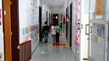 Fotografia em enquadramento amplo com ponto de fuga central de um corredor de ambiente escolar. Em primeiro plano, no centro, três crianças pequenas são vistas de costas caminhando lado a lado pelo corredor iluminado. A criança da esquerda veste casaco escuro e calça jeans; a do meio veste colete rosa claro e calça cinza; e a da direita veste casaco verde e calça escura. As paredes brancas do corredor exibem trabalhos infantis e papéis coloridos afixados em fileiras. À esquerda, há um portãozinho de madeira marrom e portas de salas de aula. À direita, vê-se uma porta de vidro decorada com recortes de corações cor-de-rosa e extintores de incêndio fixados na parede. O piso cinza possui sinalizações coloridas e setas direcionais verdes.