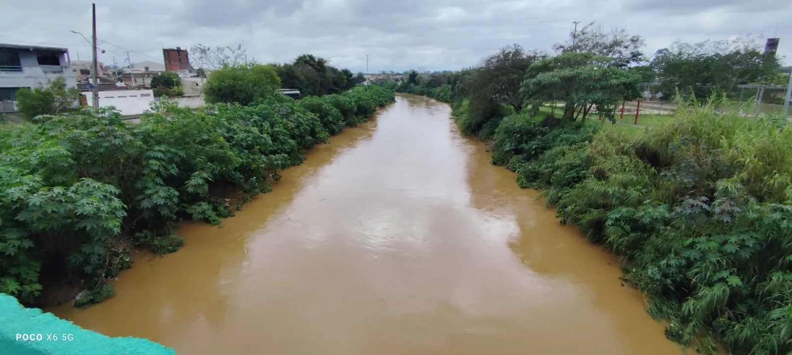 A foto mostra o Rio Barigui cheio, com água amarelada e encostas verdes preservadas, passando em meio a duas ruas do bairro CIC.