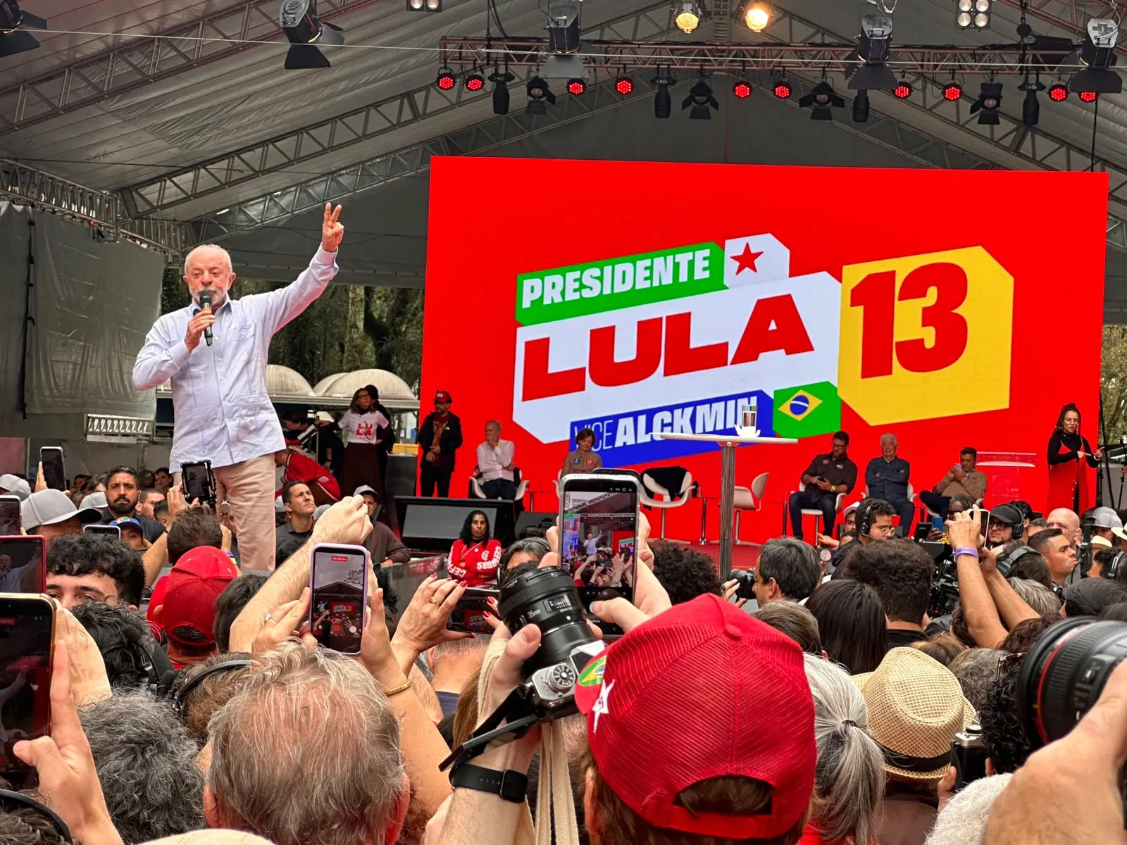 Foto mostra Lula de camisa branca e caça bege, com o braço levantado e dedo em sinal de V, em frente ao público e uma placa vermelha que diz 'Lula 13'