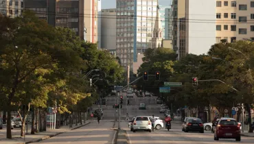 Fotografia em enquadramento amplo com profundidade de campo de uma larga avenida urbana com pouca movimentação de veículos e pedestres. A perspectiva segue em aclive rumo ao centro da imagem. Em primeiro plano, no asfalto claro iluminado pela luz suave do final da tarde, trafegam poucos carros, um pedestre atravessa a via e um motociclista carrega uma mochila de entrega vermelha à direita. Ladeando a pista, fileiras de árvores com copas verdes e densas cobrem as calçadas. Ao fundo, erguem-se diversos edifícios residenciais e comerciais de diferentes alturas e estilos arquitetônicos, incluindo a torre de uma igreja de estilo neogótico em tom claro no horizonte sob céu limpo.