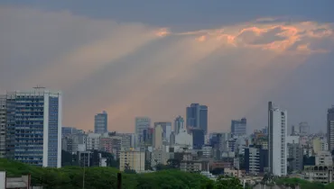 Fotografia em enquadramento plano geral e panorâmico da linha do horizonte urbano (skyline) de uma cidade. Em primeiro plano, no canto inferior, estende-se uma faixa de copa de árvores verdes e densas sob o nível das edificações. No plano intermediário, alinham-se diversos prédios e edifícios comerciais e residenciais de variadas alturas, formatos e cores. Ao fundo, o céu acinzentado de fim de tarde é cortado por feixes de luz solar alaranjada (rayos crepusculares) projetados a partir de brechas nas nuvens densas na parte superior da imagem.