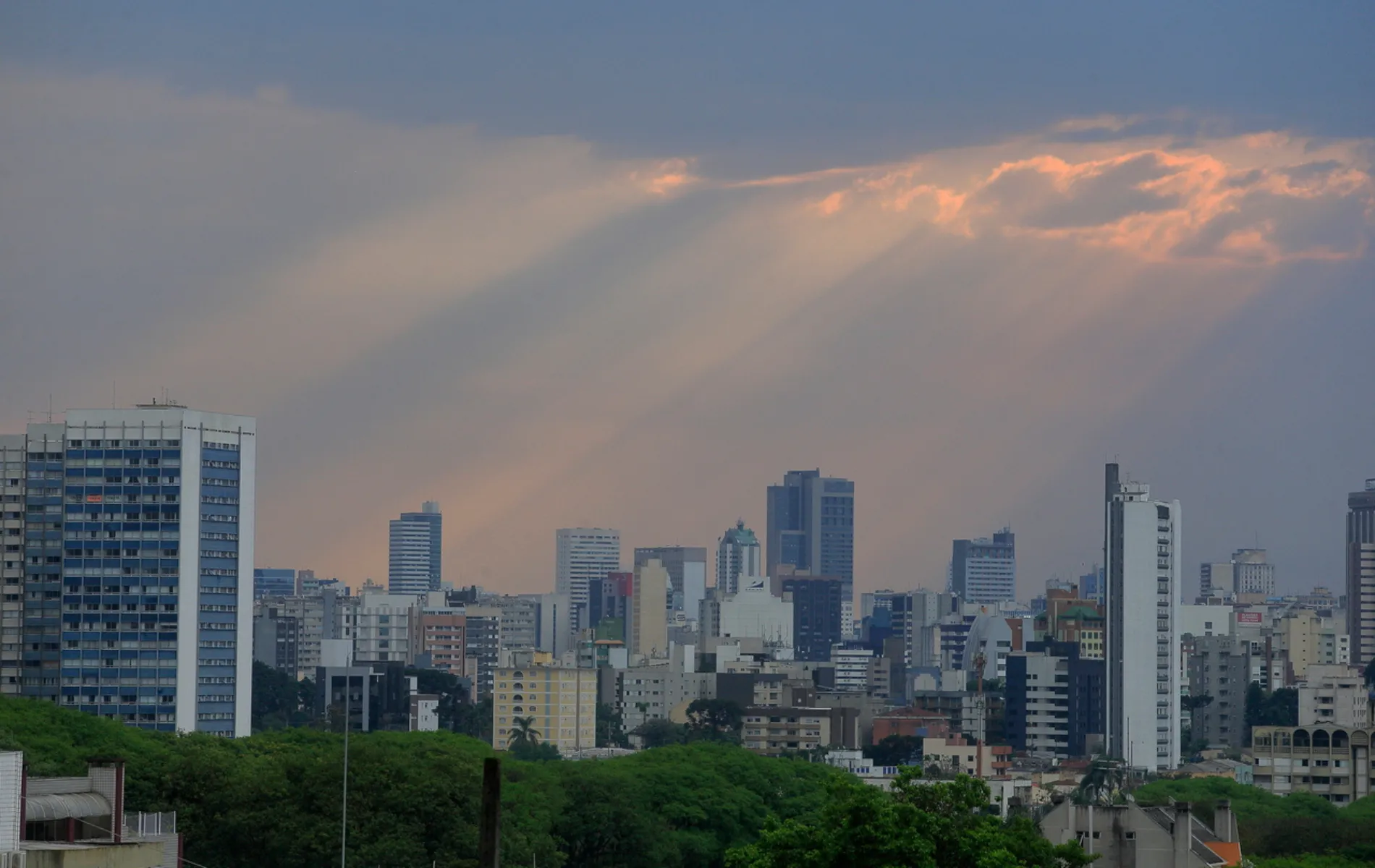 Fotografia em enquadramento plano geral e panorâmico da linha do horizonte urbano (skyline) de uma cidade. Em primeiro plano, no canto inferior, estende-se uma faixa de copa de árvores verdes e densas sob o nível das edificações. No plano intermediário, alinham-se diversos prédios e edifícios comerciais e residenciais de variadas alturas, formatos e cores. Ao fundo, o céu acinzentado de fim de tarde é cortado por feixes de luz solar alaranjada (rayos crepusculares) projetados a partir de brechas nas nuvens densas na parte superior da imagem.