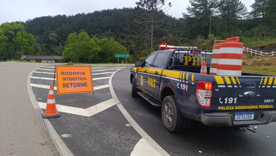 Fotografia em enquadramento médio e ângulo oblíquo de um ponto de bloqueio viário em uma rodovia. À direita, em primeiro plano, está a traseira de uma caminhonete da Polícia Rodoviária Federal (PRF), na cor azul-escura com detalhes em amarelo, giroflex ligado e inscrição "POLÍCIA RODOVIÁRIA FEDERAL", acompanhada do número de emergência "191" e placa "SEY1I86". Na caçamba do veículo há barris e cones de sinalização. À esquerda, sobre o asfalto pintado com marcas de canalização zebradas, há uma placa de sinalização de trânsito alaranjada com a mensagem em caixa alta "RODOVIA INTERDITADA RETORNE", ladeada por um cone laranja e branco. Ao fundo, veem-se morros cobertos por vegetação densa e pinheiros (araucárias) sob céu nublado.