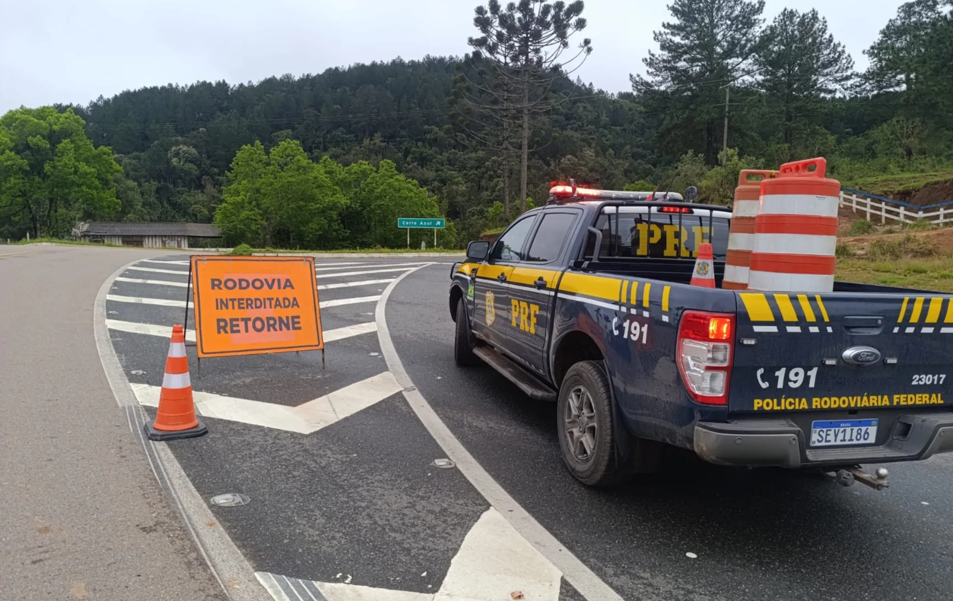 Fotografia em enquadramento médio e ângulo oblíquo de um ponto de bloqueio viário em uma rodovia. À direita, em primeiro plano, está a traseira de uma caminhonete da Polícia Rodoviária Federal (PRF), na cor azul-escura com detalhes em amarelo, giroflex ligado e inscrição "POLÍCIA RODOVIÁRIA FEDERAL", acompanhada do número de emergência "191" e placa "SEY1I86". Na caçamba do veículo há barris e cones de sinalização. À esquerda, sobre o asfalto pintado com marcas de canalização zebradas, há uma placa de sinalização de trânsito alaranjada com a mensagem em caixa alta "RODOVIA INTERDITADA RETORNE", ladeada por um cone laranja e branco. Ao fundo, veem-se morros cobertos por vegetação densa e pinheiros (araucárias) sob céu nublado.