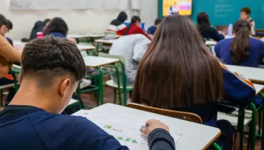 Fotografia em enquadramento amplo com plano de profundidade tomada a partir do fundo de uma sala de aula de escola ou universidade. Em primeiro plano, à esquerda, vê-se a parte posterior da cabeça de um estudante de cabelos curtos castanhos e corte raspado nas laterais, curvado sobre a carteira escolar enquanto preenche uma folha de respostas ou prova. À sua direita, destaca-se uma estudante de costas, com longos cabelos castanhos lisos que cobrem suas costas. Ao fundo, o ambiente escolar se estende em perspectiva com diversas fileiras de carteiras ocupadas por outros alunos realizando a atividade. Na parede ao fundo, observa-se uma televisão ligada fixada no alto e um quadro escolar verde, além de um professor ou aplicador sentado à mesa sob iluminação fluorescente uniforme.