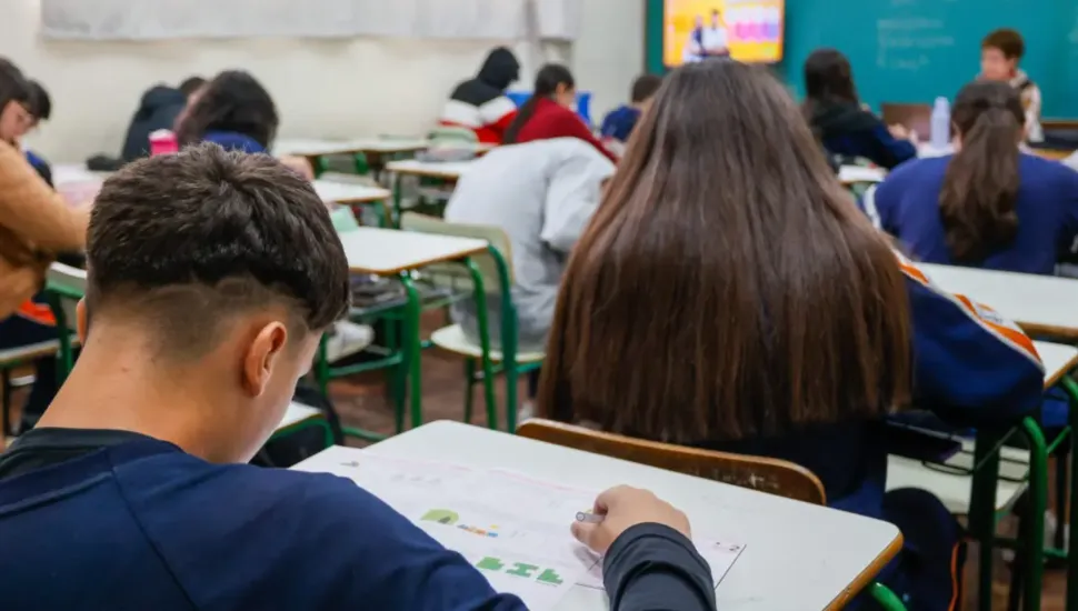 Fotografia em enquadramento amplo com plano de profundidade tomada a partir do fundo de uma sala de aula de escola ou universidade. Em primeiro plano, à esquerda, vê-se a parte posterior da cabeça de um estudante de cabelos curtos castanhos e corte raspado nas laterais, curvado sobre a carteira escolar enquanto preenche uma folha de respostas ou prova. À sua direita, destaca-se uma estudante de costas, com longos cabelos castanhos lisos que cobrem suas costas. Ao fundo, o ambiente escolar se estende em perspectiva com diversas fileiras de carteiras ocupadas por outros alunos realizando a atividade. Na parede ao fundo, observa-se uma televisão ligada fixada no alto e um quadro escolar verde, além de um professor ou aplicador sentado à mesa sob iluminação fluorescente uniforme.