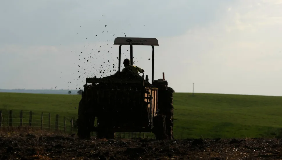 Fotografia em enquadramento médio com contra-luz de um operador conduzindo um trator agrícola em uma área rural. Visto de costas, o veículo está posicionado ao centro da composição, sob contraluz, projetando uma silhueta contra o horizonte. O maquinário espalha adubo ou terra escura para trás, lançando diversos fragmentos no ar em movimento contínuo. No primeiro plano, o solo de terra arada exibe textura escura e irregular. Ao fundo, uma vasta extensão de campo verde suavemente ondulado estende-se sob a iluminação difusa de um céu nublado ao fim da tarde.