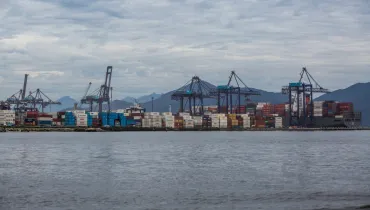 Fotografia em enquadramento amplo e vista panorâmica de um terminal portuário às margens de uma baía. Em primeiro plano, ocupa a metade inferior da imagem a superfície da água em tom cinza-azulado com pequenas ondas suaves. No plano intermediário, estende-se a faixa do porto com milhares de contêineres coloridos empilhados — predominantemente azul, vermelho, branco e amarelo — e um navio cargueiro atracado à direita. Diversos guindastes e portêineres de grande porte erguem-se ao longo da extensão do cais. Ao fundo, a paisagem é delimitada pela silhueta de montanhas sob um céu totalmente coberto por nuvens cinzentas e densas em iluminação difusa diurna.